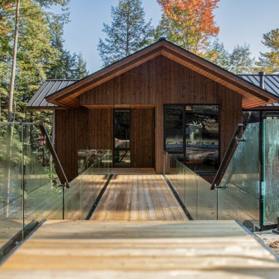 Modern lakeside cabin exterior designed by Alderstone Homes, featuring a glass bridge walkway and natural wood façade surrounded by forest.