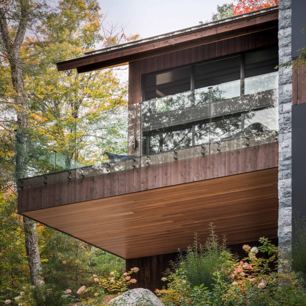 Elevated deck with glass railing and natural wood siding designed by Alderstone Homes, surrounded by forest views.